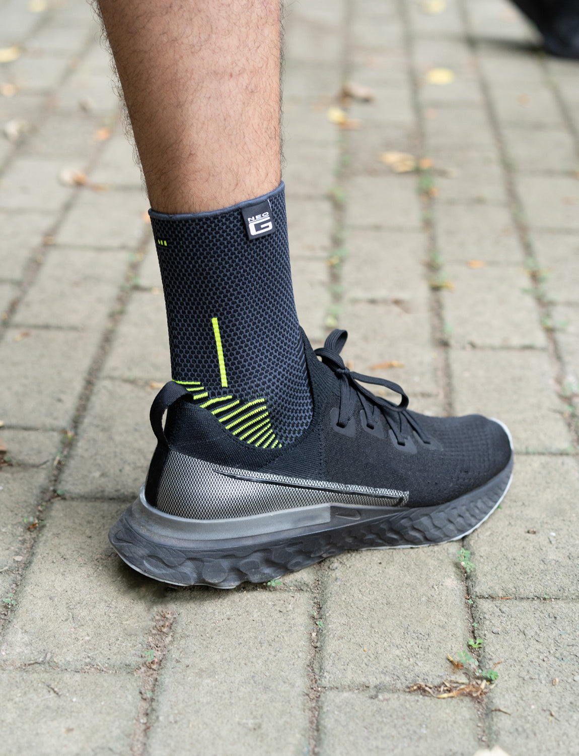 A close-up of a person's lower leg and foot wearing a Neo G USA Active Ankle Support with bright green detailing and breathable fabric, on a cobblestone path with scattered leaves.
