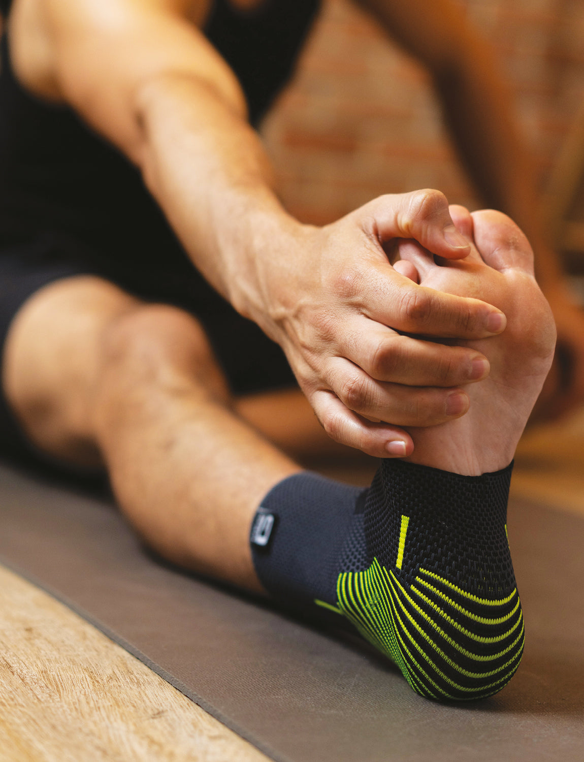 A person sitting on a yoga mat holding their foot, wearing a black and green sock with multi-zone compression, focusing on stretching or inspecting their foot, in a room with a brick wall background while using Neo G USA Active Ankle Support.