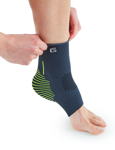 A person adjusting a Neo G USA Active Ankle Support with multi-zone compression on their left leg, shown against a white background. The brace features a combination of blue and neon yellow colors.