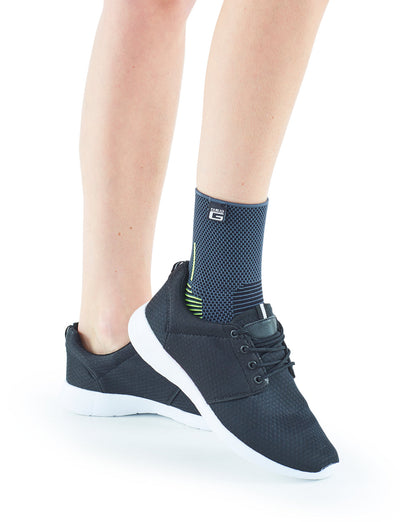 A close-up of a person's legs showing one foot wearing a stylish black sneaker and a Neo G USA Active Ankle Support featuring multi-zone compression. The background is plain white.